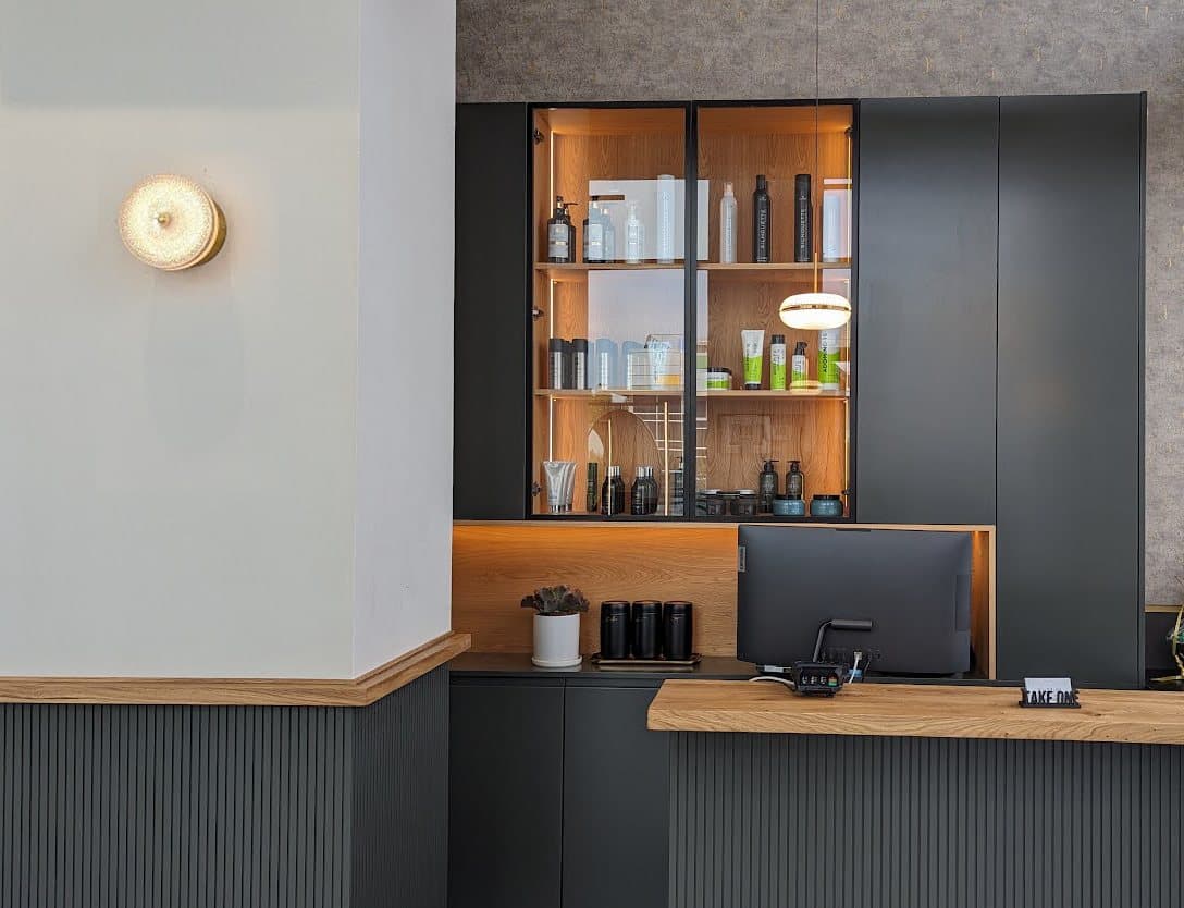 דלפק קבלה מעץ במספרה, עם ויטרינת מוצרים מוארת, חיפוי מחורץ כהה ומנורת קיר עגולה