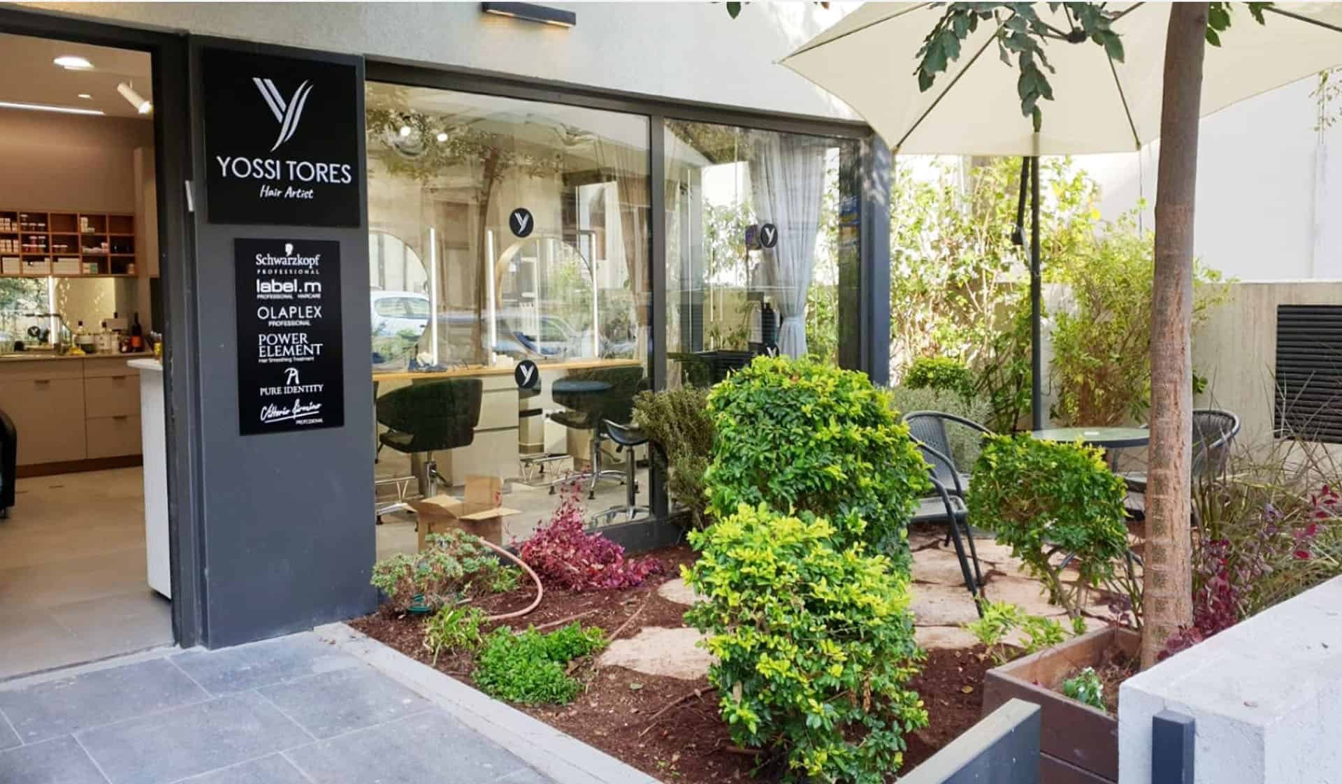 חזית מספרת יוסי טורס בתל אביב — שלט עם הלוגו, קיר זכוכית וגינה מטופחת עם שמשייה